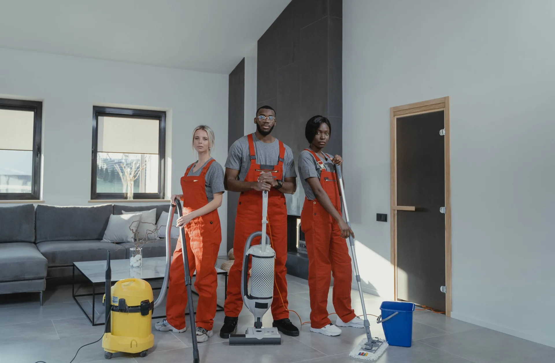 Three cleaning professionals with vacuum cleaners and a mop in a bright living room